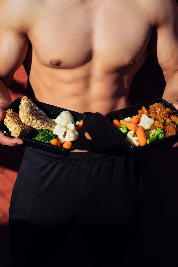  A shirtless man holding two trays of healthy meal prep food.